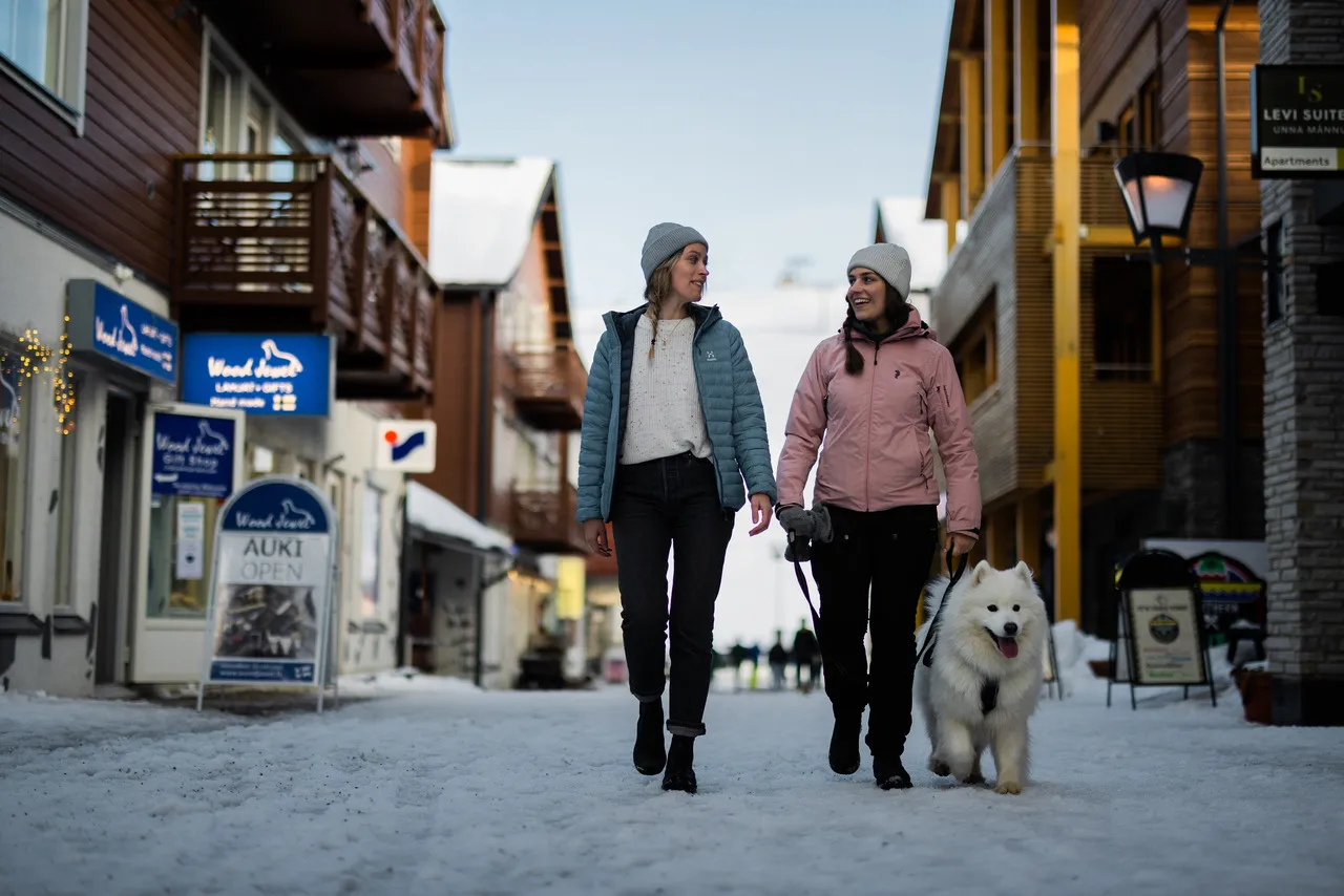 Two women walking a dog along a snowy shopping street in Levi village